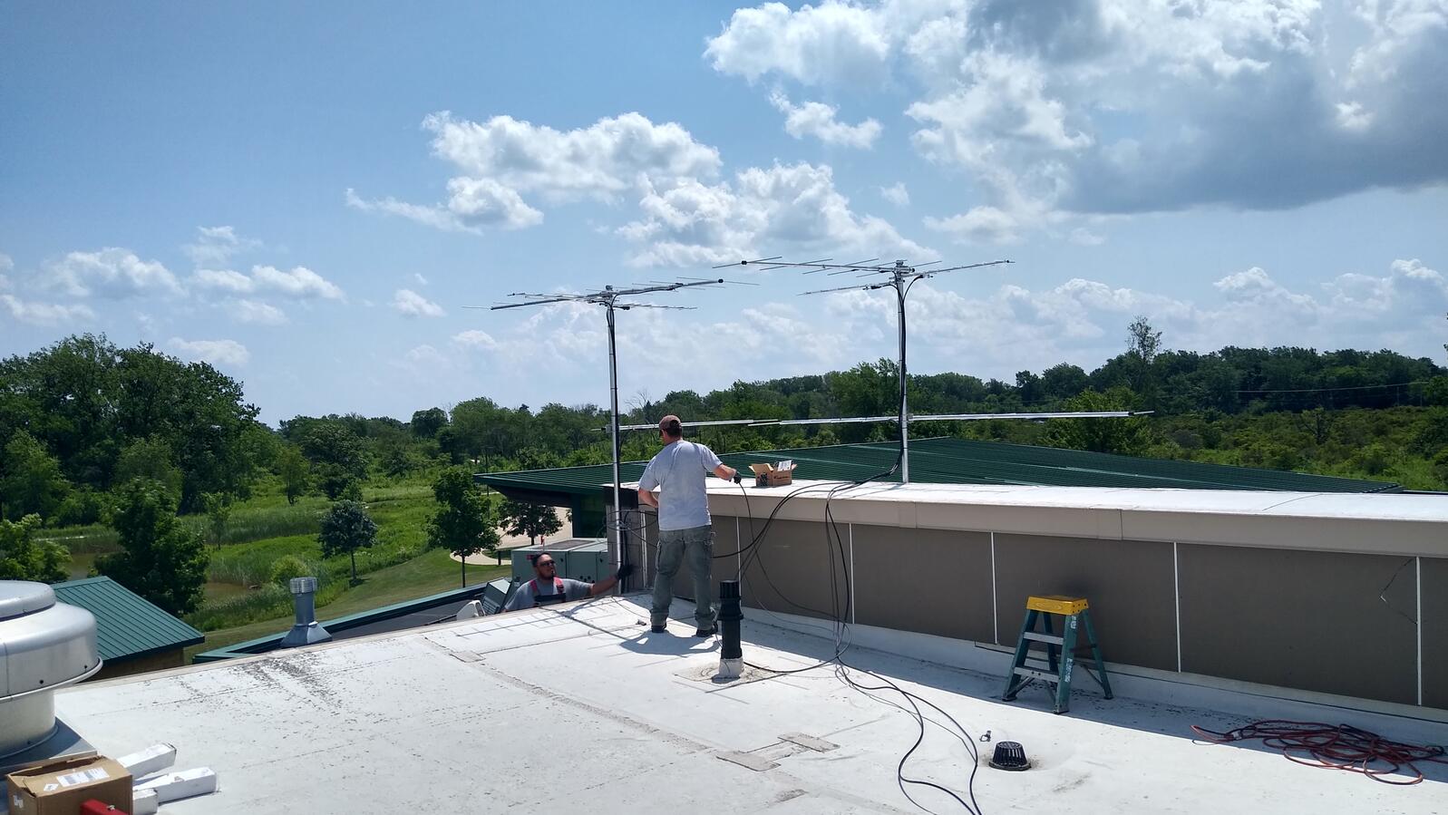 Two sets of antennae from the Greenbelt Forest Preserve Motus station sit on a rooftop with green trees, blue sky and white clouds in the background.  